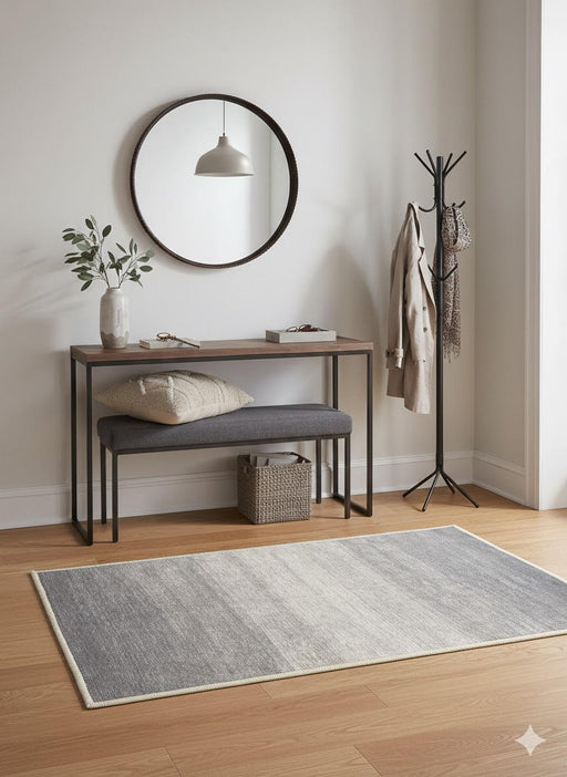 2' x 3' Gray Ivory / Cream Ombre Washable Area Rug placed / styled in a minimalist entryway beneath a wood and metal console table with a cushioned bench, round wall mirror, and black coat rack.