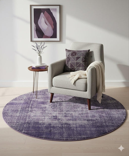 5' Round Purple Plum Soft Gray Round Heathered Abstract/Distressed Modern area rug placed in a bright living room, styled beneath a light gray upholstered accent chair with a geometric plum pillow, a small wooden side table with decorative vase, framed abstract wall art, and soft natural light streaming across pale wood floors.