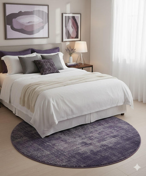 5' Round Purple Plum Soft Gray Round Heathered Abstract/Distressed Modern area rug placed in a serene bedroom, styled partially under a neatly made bed with white bedding, layered accent pillows in gray and plum tones, matching abstract wall art, a bedside table with lamp, and sheer curtains filtering soft daylight.