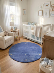 5' round denim blue & soft gold accent minimal motif modern area rug styled in a soft nursery beside a white crib, armchair, and framed animal artwork.