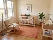 5' x 7' Rectangular Peach Coral & Rust Solid Textured Border Modern Area Rug styled in a nursery with a wooden crib and cream rocking chair.