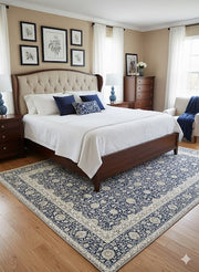 5' x 7' Rectangular blue ivory soft gray beige traditional floral area rug placed in a serene bedroom with a tufted upholstered bed, white bedding, wooden nightstands, decorative pillows, framed botanical artwork, and warm wood flooring illuminated by soft daylight.