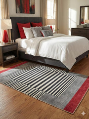 5' x 7' black, ivory, and red modern striped rectangular area rug placed in a bright bedroom beneath a dark upholstered bed with white bedding and warm wood flooring.