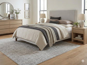 5' x 7' soft gray ivory washable heathered solid area rug placed in a calming bedroom with layered neutral bedding, light wood furniture, and soft natural light.