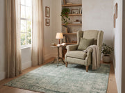 5' x 8' Rectangular Sage Green & Ivory Distressed Floral Ornamental Border Vintage Transitional Area Rug styled in a cozy reading nook with a beige wingback armchair and wooden side table.