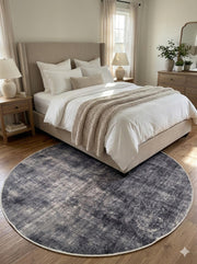 6' round charcoal ivory light beige distressed modern area rug placed under a bed in a bright neutral bedroom.