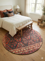 6' round deep rust red and charcoal black southwestern geometric area rug styled at the foot of a bed in a warm, neutral bedroom.