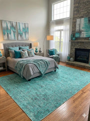 6' x 9' teal blue vintage distressed washable area rug placed beneath a modern bed with teal bedding accents in a spacious bedroom with a stone fireplace.