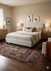 7' x 10' charcoal and burnt orange tribal geometric area rug styled partially under a bed in a modern bedroom with beige bedding and wooden furniture.