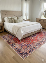 7' x 10' red, navy blue, and cream floral medallion traditional area rug positioned under a bed in a cozy bedroom with light wood furniture.