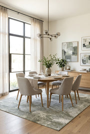7' x 7' Square Light Gray & Sage Green Distressed Abstract Modern Washable Area Rug styled in a dining room with a wooden dining table and beige upholstered chairs.