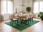 7' x 9' Rectangular Emerald Green & Black Distressed Abstract Modern Washable Area Rug styled in a dining room beneath a wooden dining table and cream upholstered chairs.