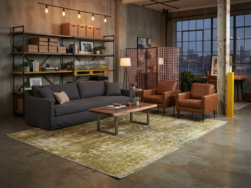 7' x 9' Rectangular Olive Gold Beige Distressed Abstract Vintage Modern Washable Area Rug styled in a living room with a charcoal gray sofa, brown leather armchairs, and wooden coffee table.