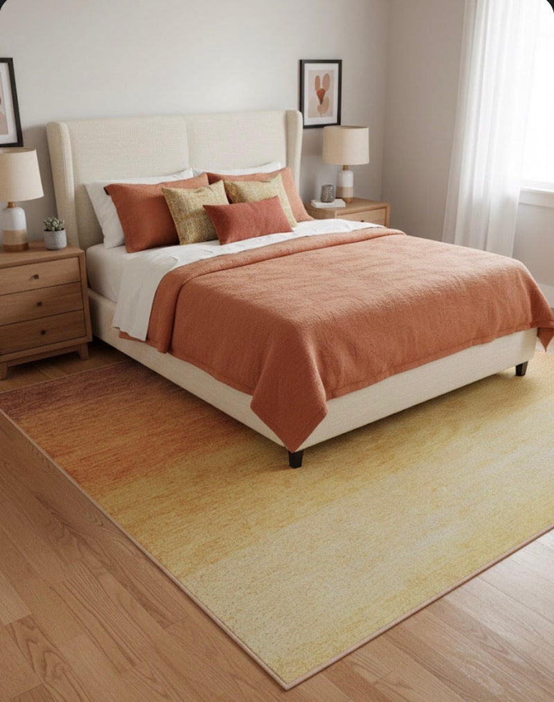 7' x 9' golden yellow and warm orange ombre washable area rug placed beneath a bed in a bright bedroom with soft neutral furniture and warm-toned bedding.