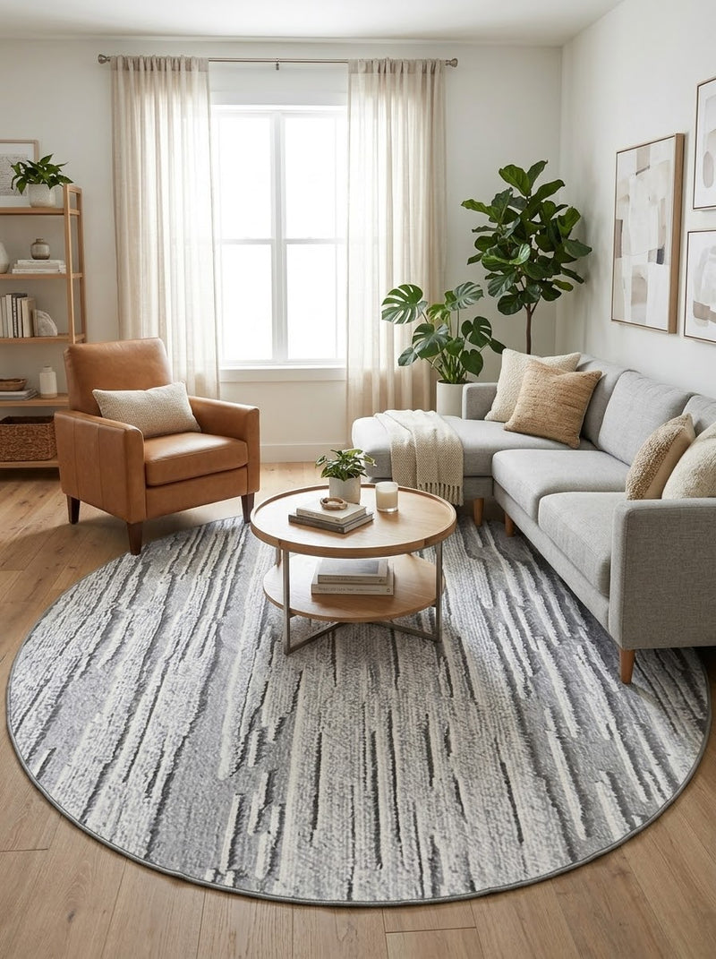 8' Round Gray Ivory Abstract Striped Modern Washable Area Rug styled in a living room with a gray sectional sofa, tan armchair and round coffee table.
