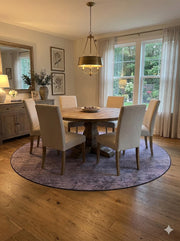 8' round taupe distressed modern rug placed under a rustic round dining table with upholstered chairs in a bright neutral dining space.