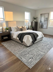 8' x 10' gray geometric modern rug placed under a neutral upholstered bed with layered white bedding in a calm contemporary bedroom.