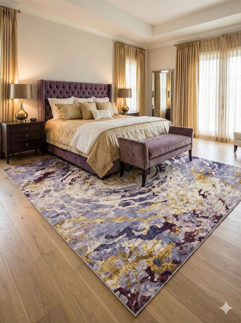 8' x 10' purple, gold, and ivory washable abstract floral area rug placed beneath a bed in a luxurious bedroom featuring a tufted purple headboard and warm neutral bedding.