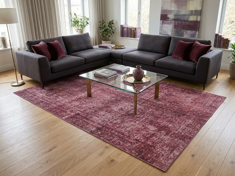 8' x 12' Rectangular Rose Pink Burgundy Distressed Abstract Vintage Area Rug styled in a modern living room with a charcoal gray sectional sofa, burgundy accent pillows, glass coffee table, and large windows.